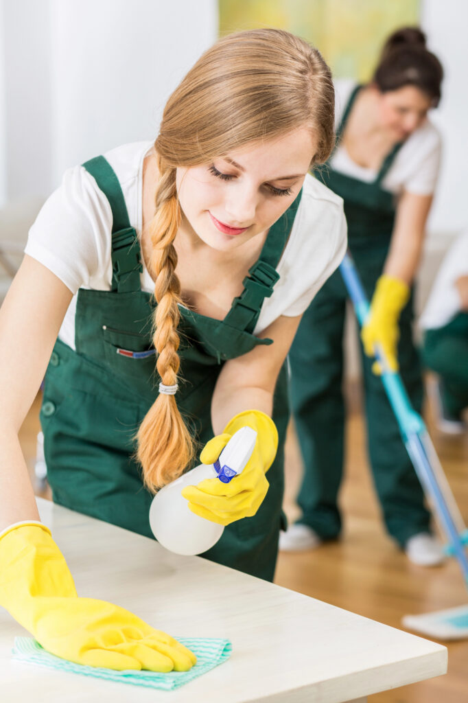 Professional cleaner in green overalls and yellow gloves wiping a table surface with a cloth and spray bottle in a residential or Airbnb property